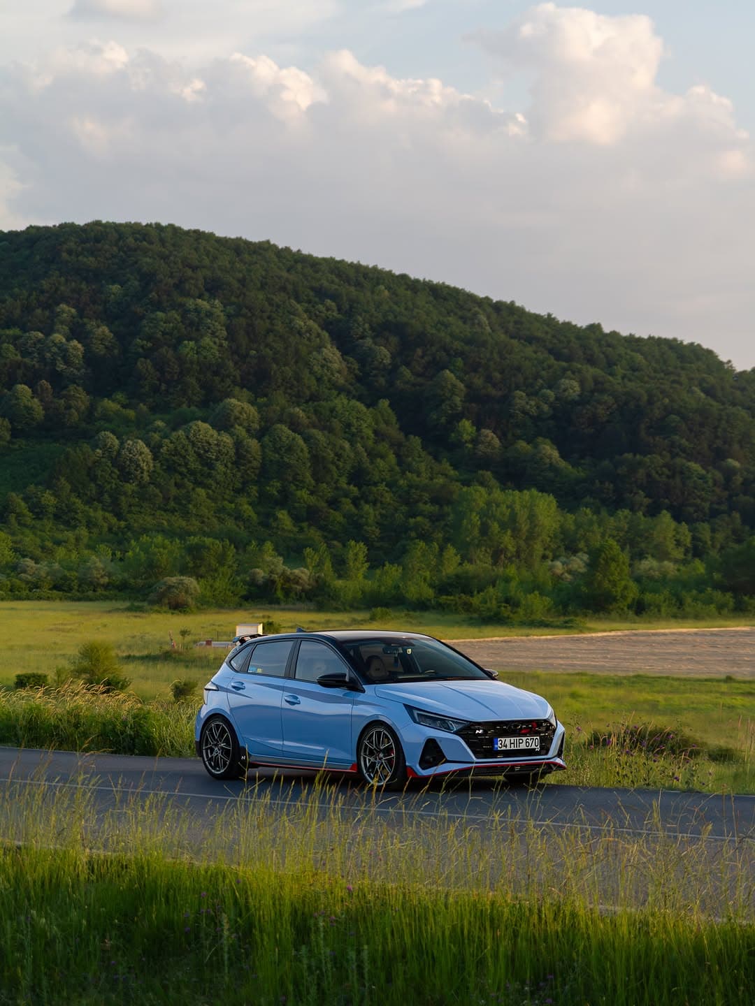 A blue Hyundai i20 N parked on a country road with a green hillside in the background.