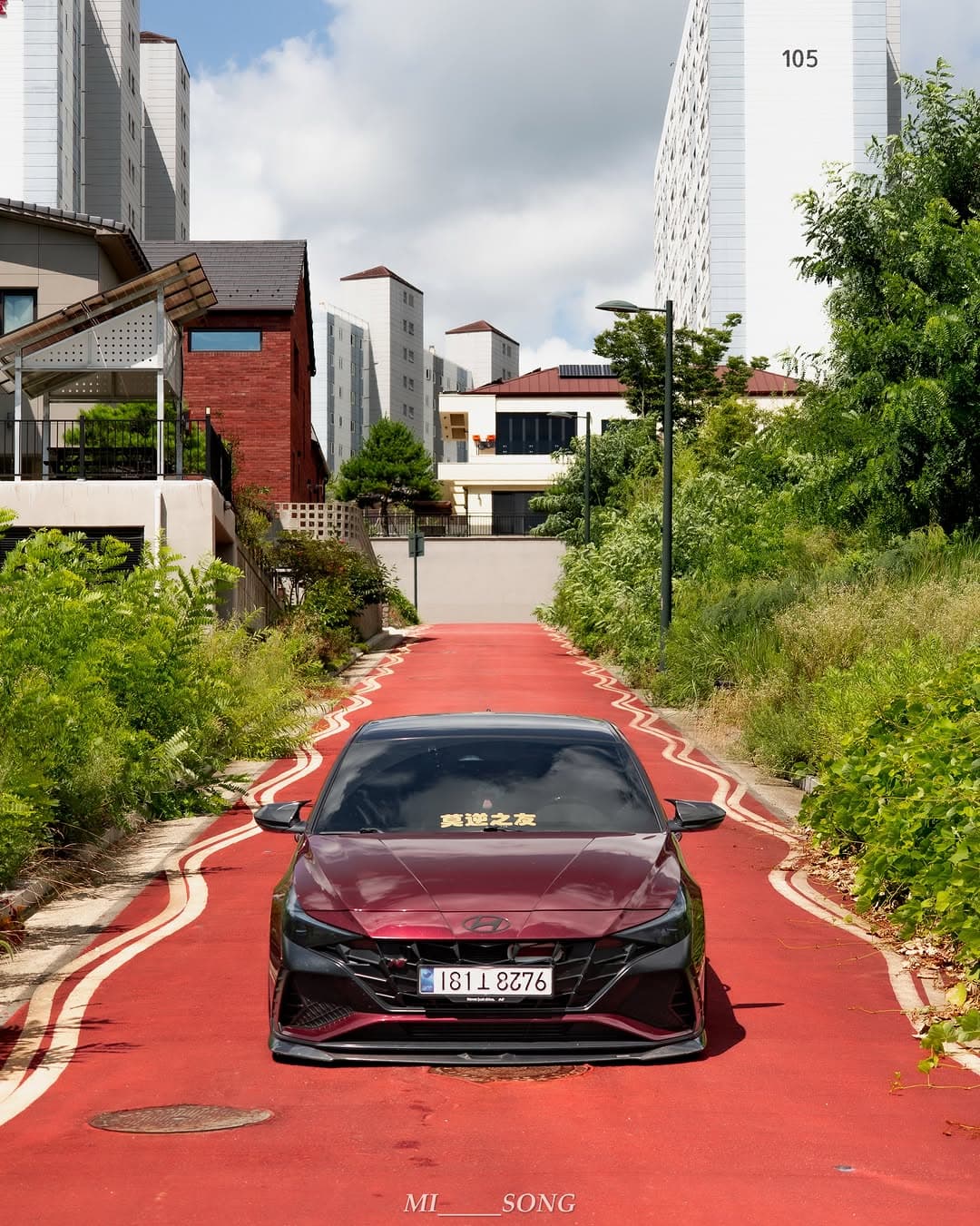 A burgundy Hyundai Elantra N parked on a red road between greenery and apartment buildings.