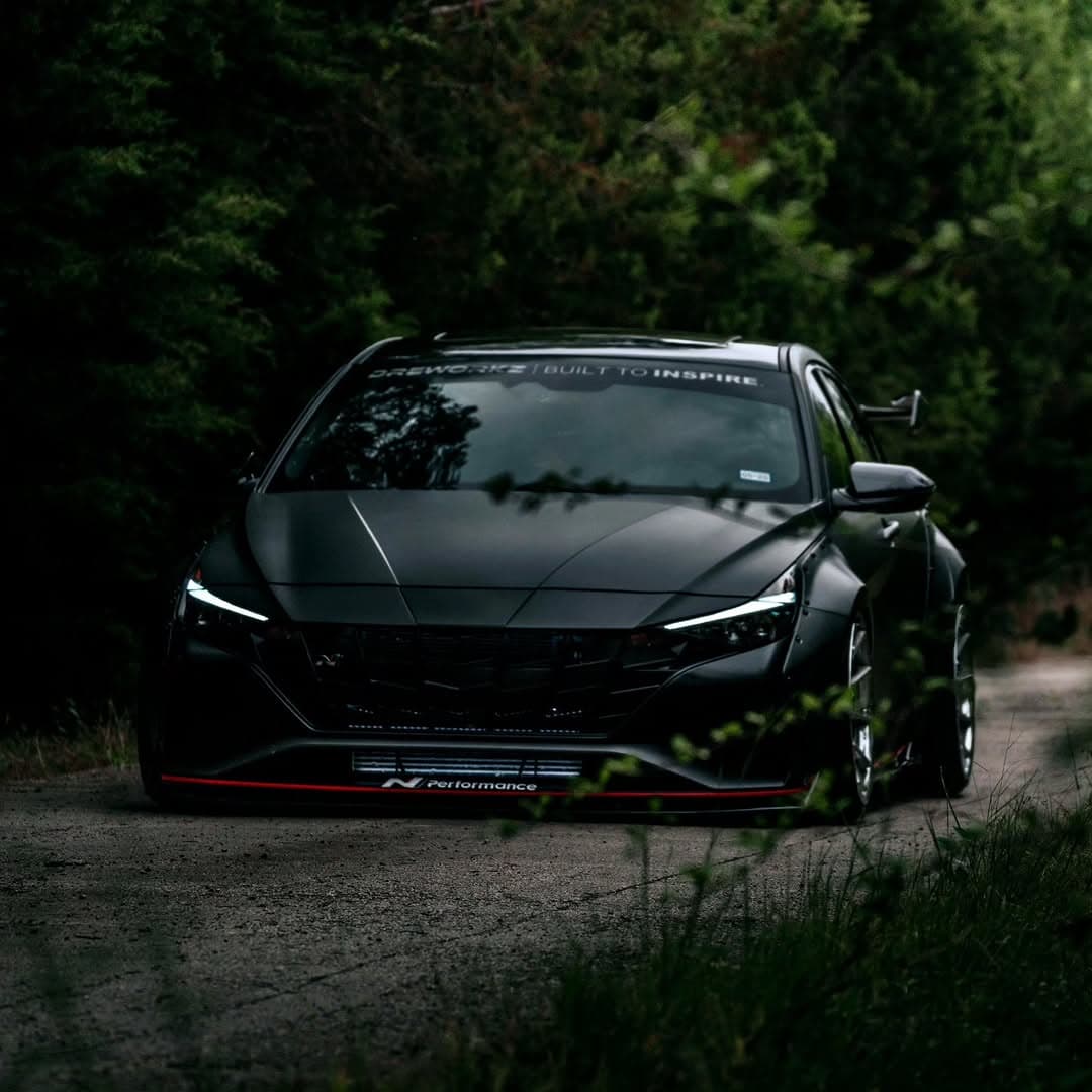 A matte black widebody Hyundai Elantra N photographed head-on on a narrow road surrounded by trees.